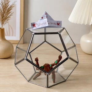 May include: A geometric glass terrarium with a silver metal frame containing a brown and green articulated figure. A white paper hat sits on top of the glass, featuring red stars and the handwritten text, Savior of the Universe.