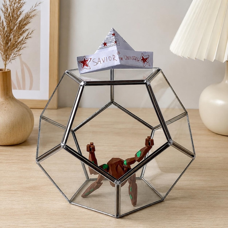 May include: A geometric glass terrarium with a silver metal frame containing a brown and green articulated figure. A white paper hat sits on top of the glass, featuring red stars and the handwritten text, Savior of the Universe.
