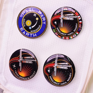 May include: Four circular pin badges with space-themed designs. One badge features the text "HAIL MARY" and a blue and gold design. The other three badges have a rocket ship design with the text "PROJECT HAIL MARY" and "TEAM GRACE & ROCKY".