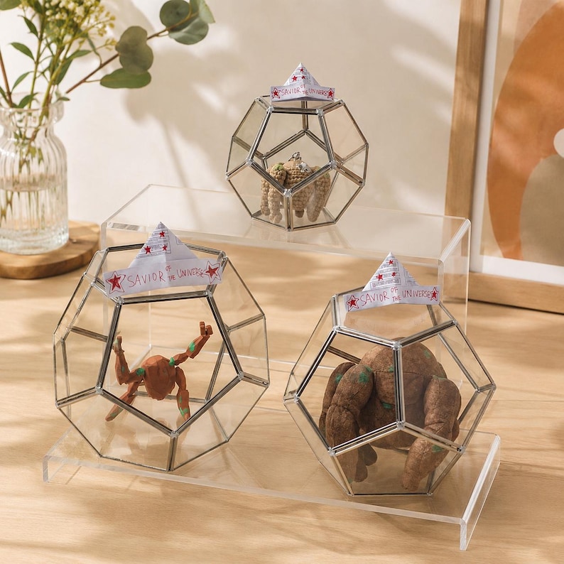 May include: Three geometric glass terrariums with metal frames, each containing a small figurine. Each terrarium is topped with a paper boat ornament featuring the text "SAVIOR OF THE UNIVERSE" with red stars on both ends of the paper banner.