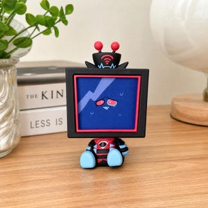 Puede incluir: Una peque&ntilde;a figura coleccionable con una cabeza en forma de televisor negro, un marco rojo y una pantalla azul que muestra un rostro de personaje estresado. La figura tiene dos antenas rojas en la parte superior y un cuerpo negro y rojo con extremidades azul claro.