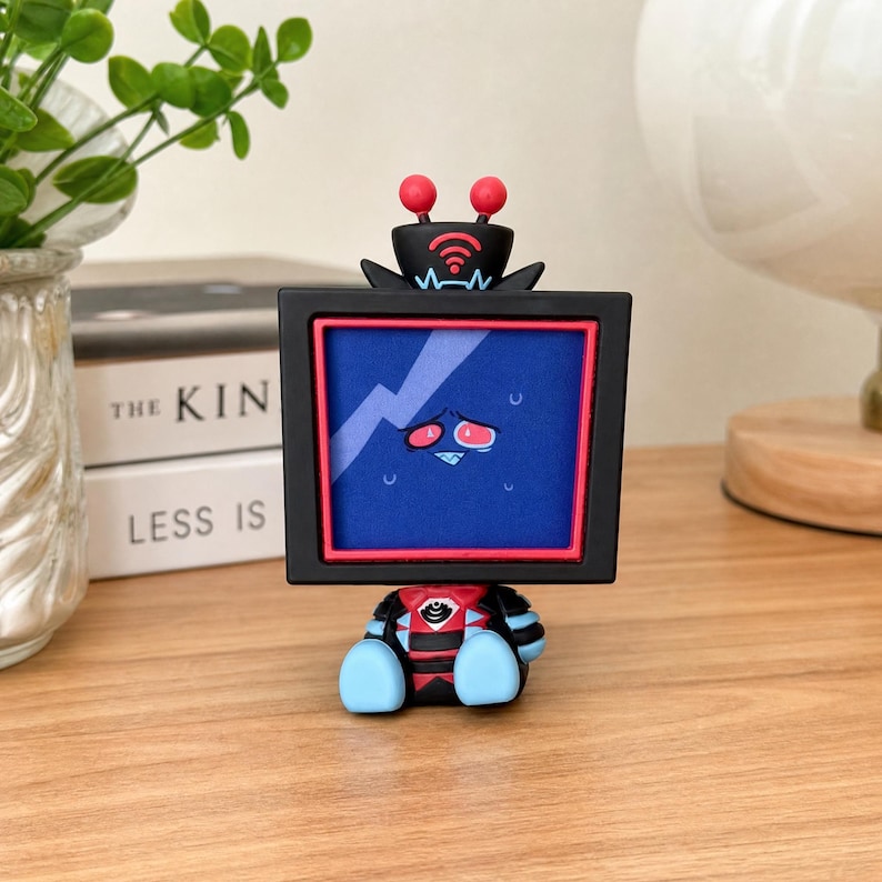 Puede incluir: Una peque&ntilde;a figura coleccionable con una cabeza en forma de televisor negro, un marco rojo y una pantalla azul que muestra un rostro de personaje estresado. La figura tiene dos antenas rojas en la parte superior y un cuerpo negro y rojo con extremidades azul claro.