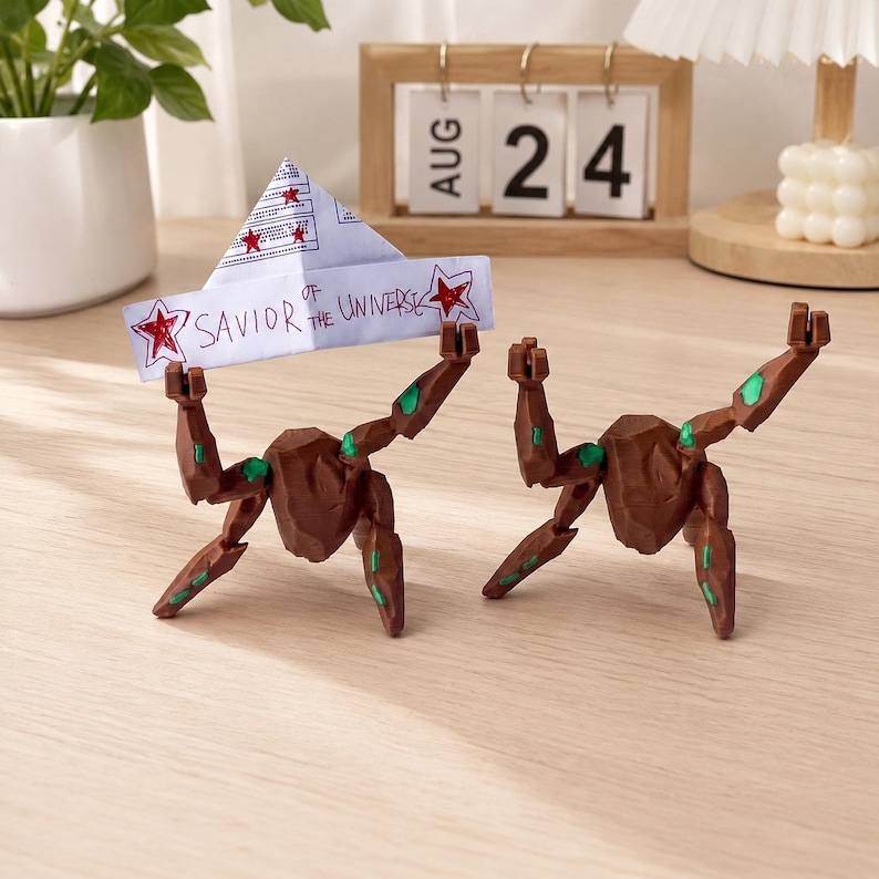 May include: Two brown, rock-like articulated figures with green accents on their limbs. One figure holds a white paper boat with red star drawings and the handwritten text "SAVIOR OF THE UNIVERSE" in black ink. The figures have multiple joints.