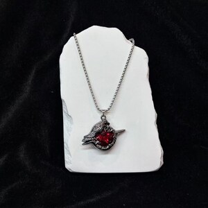 May include: A silver-toned necklace with a pendant featuring a dark metal bird design holding a large, round, red gemstone. The necklace is displayed on a white, textured surface against a black background.