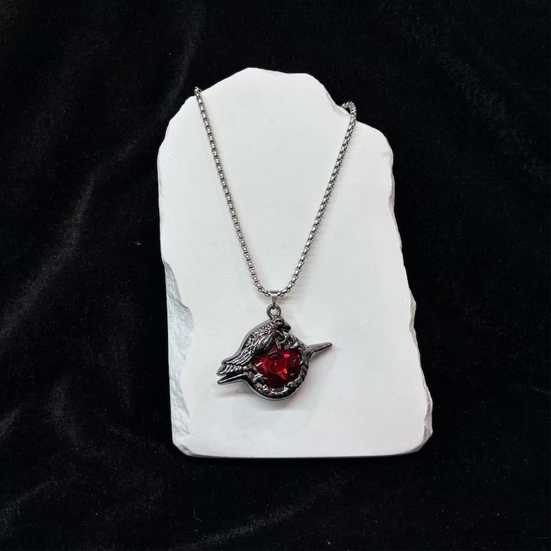 May include: A silver-toned necklace with a pendant featuring a dark metal bird design holding a large, round, red gemstone. The necklace is displayed on a white, textured surface against a black background.