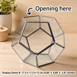 May include: A clear glass geometric terrarium with a pentagonal frame and metallic edges. The object is labeled Display Dome B and measures 6.89 x 6.89 x 5.91 inches. An arrow points to the top opening with the text Opening here.