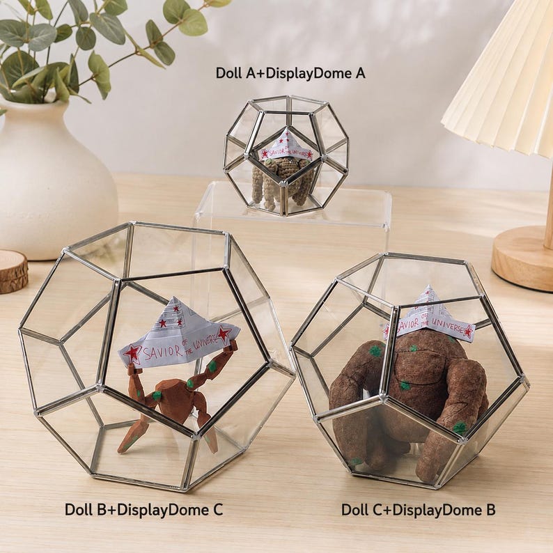 May include: Three geometric glass display domes with silver metal frames. One dome contains a small brown crocheted doll, while the other two contain brown dolls wearing paper hats labeled Savior of the Universe with red star accents.