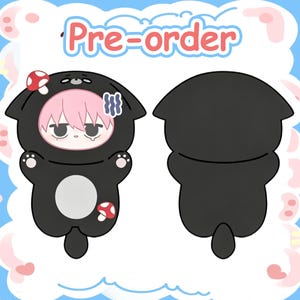 May include: A plush toy character wearing a black animal hood with cat ears, a pink mushroom on the left ear, and a red mushroom on the lower body. The character has pink hair, a tired expression, and a white belly patch. Text reads Pre-order.