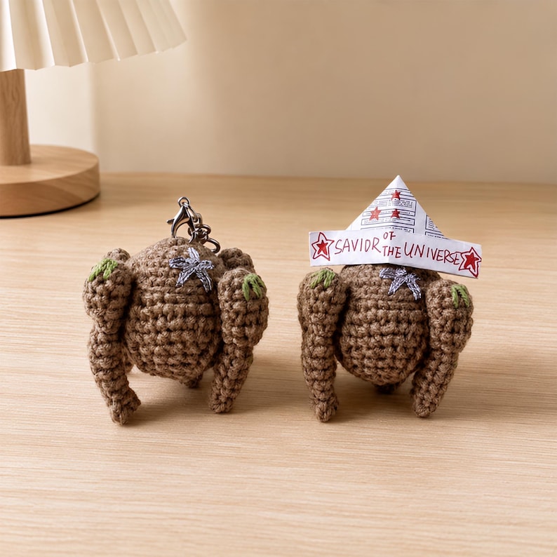 May include: Two brown crocheted chicken figures with small green accents on the wings. One figure features a metal keychain attachment. The other figure wears a white paper hat with red stars and the handwritten text SAVIOR OF THE UNIVERSE.