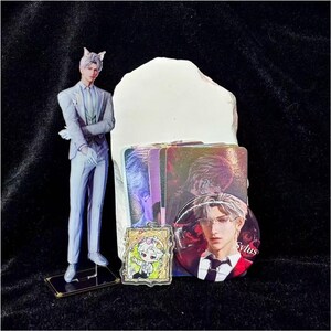 May include: A collection of anime-themed merchandise, including a figure of a man in a gray suit with cat ears, a button with the name "Sylus", and several holographic cards. A small, framed cartoon character is also present.
