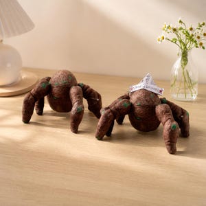 May include: Two brown, textured, spider-like plush figures with green spots on their legs and bodies. One figure wears a small, white, triangular paper hat with the text "SAVIOR OF THE UNIVERSE" printed on it in black ink with red stars.
