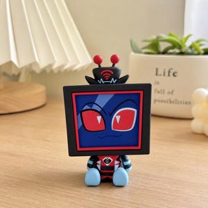 Puede incluir: Una peque&ntilde;a figura coleccionable con cabeza cuadrada en forma de televisi&oacute;n, pantalla azul oscuro, ojos rojos y borde rojo. La figura tiene dos antenas rojas en la parte superior, un torso negro y rojo y pies redondeados azul claro sentados.