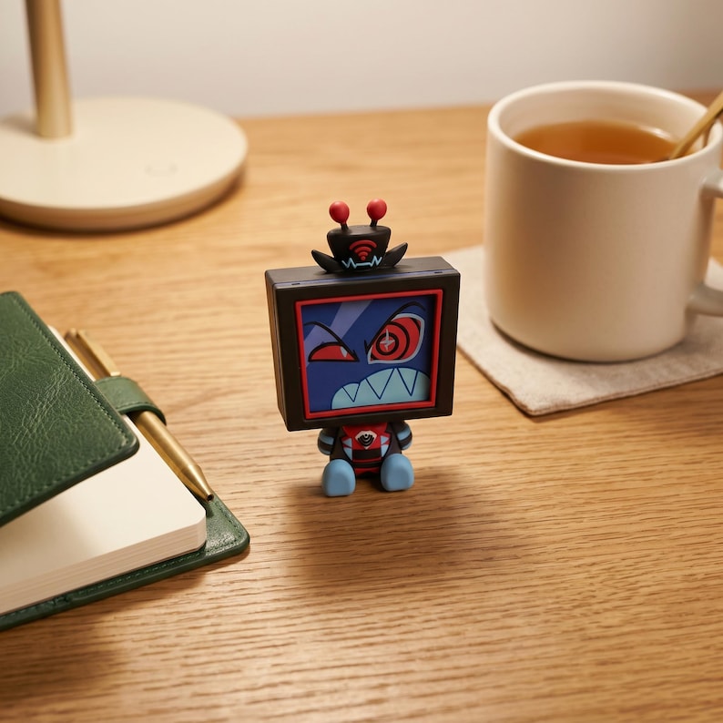 Puede incluir: Una peque&ntilde;a figura de robot de forma cuadrada con un marco negro y una pantalla que muestra una cara de dibujos animados con ojos rojos, una boca con dientes afilados y dos antenas rojas en la parte superior. El robot tiene un torso negro y pies azul claro.