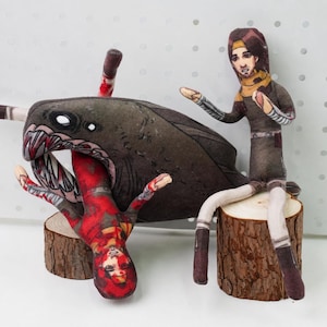 May include: Two fabric dolls featuring printed character designs. One doll is a brown-haired figure in a dark outfit, while the other is a red-toned figure being partially consumed by a large, dark, toothy creature with white eyes.