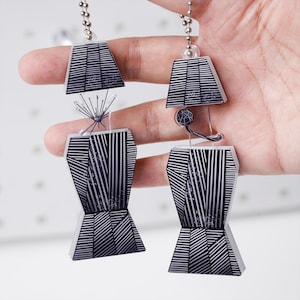 Könnte beinhalten: Zwei Schlüsselanhänger mit schwarz-weißem geometrischem Design. Jeder Schlüsselanhänger hat ein stilisiertes, abstraktes Design mit einer Kombination aus geraden Linien und Formen. Die Schlüsselanhänger sind an einer silbernen Kugelkette befestigt.