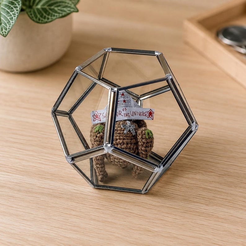 May include: A geometric glass terrarium with a silver metal frame containing a small brown crocheted figure. The figure wears a white paper crown with red stars and the handwritten text "SAVIOR OF THE UNIVERSE" in black ink.