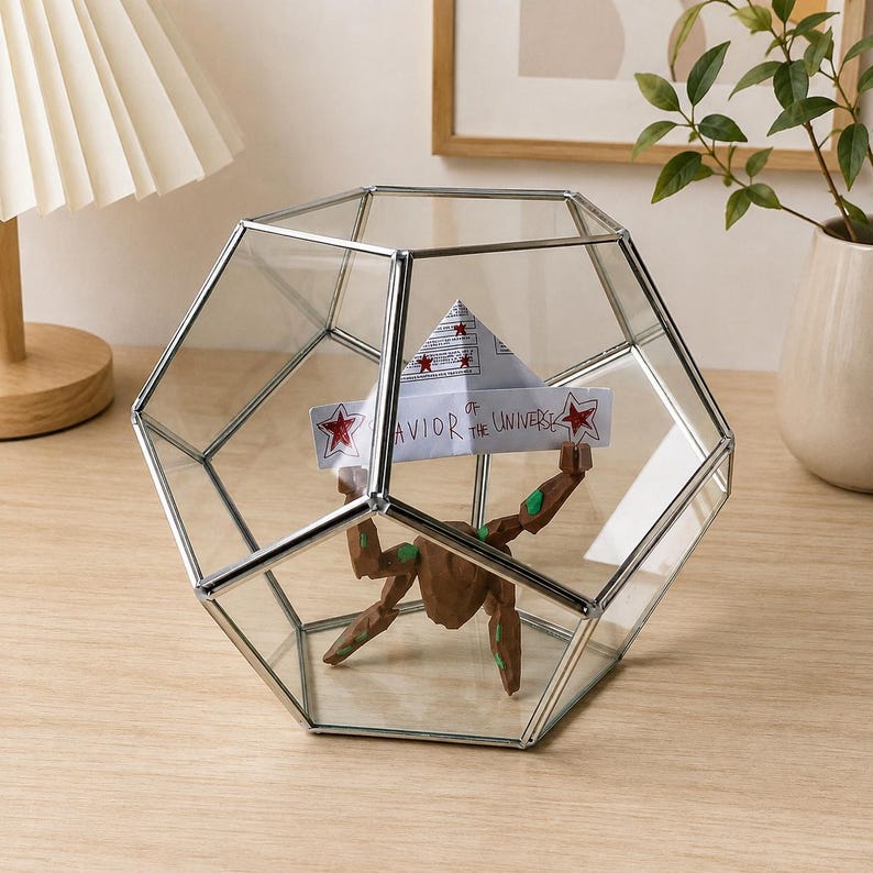 May include: A geometric glass terrarium with a silver metal frame containing a brown, multi-limbed figurine holding a white paper boat. The boat features red star illustrations and the handwritten text "SAVIOR of the UNIVERSE" in black ink.