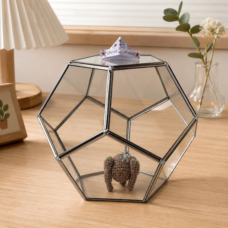 May include: A dodecahedron-shaped glass terrarium with silver metal framing. Inside sits a small brown crocheted figure. A folded paper fortune teller with the text "SAVIOR OF THE UNIVERSE" and red star accents rests on the top glass panel.