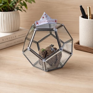 May include: A geometric glass terrarium with silver metal edges containing a small brown crocheted figure. A white paper boat sits on top of the glass structure with the handwritten text Savior of the Universe and red star illustrations.