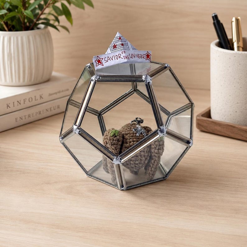 May include: A geometric glass terrarium with silver metal edges containing a small brown crocheted figure. A white paper boat sits on top of the glass structure with the handwritten text Savior of the Universe and red star illustrations.