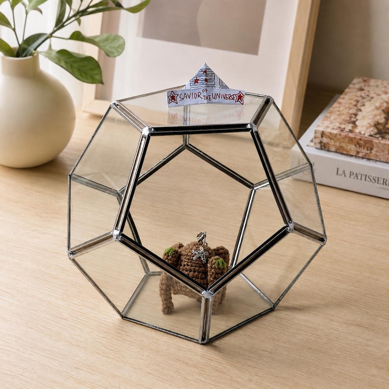 May include: A geometric glass terrarium with dark metal edges containing a small brown crocheted creature. A paper hat sits on top with the text SAVIOR OF THE UNIVERSE and red star illustrations. The structure is a dodecahedron shape with clear panels.
