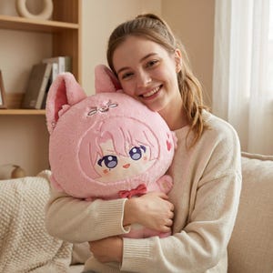 May include: A plush toy featuring a character with pink hair and large purple eyes, wearing a fuzzy pink animal hood with tall ears. The character has a small red bow tie at the neck and a stitched face with a smiling expression and pink blush marks.