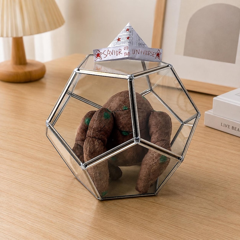 May include: A geometric glass terrarium with a silver metal frame containing a brown plush creature with green spots. A paper boat sits on top of the glass, featuring the text "SAVIOR OF THE UNIVERSE" and red star illustrations on each side.