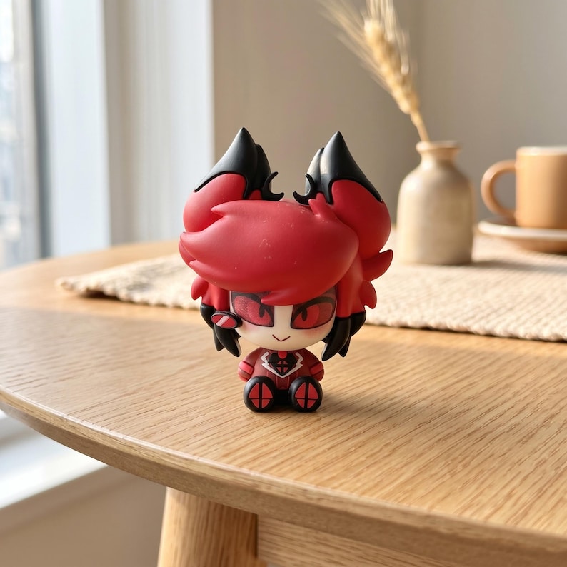 Puede incluir: Una peque&ntilde;a figura de vinilo estilizada que representa a un personaje con cabello rojo vibrante, mechones negros en forma de orejas y grandes ojos rojos. La figura tiene un rostro p&aacute;lido, una sonrisa sencilla y viste un traje rojo y negro.