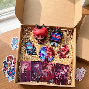 Puede incluir: Una caja de regalo de cart&oacute;n llena de art&iacute;culos de Hazbin Hotel, incluyendo dos figuras de personajes, dos llaveros, un pin circular y cuatro tarjetas art&iacute;sticas rectangulares. Varias pegatinas sueltas est&aacute;n esparcidas alrededor.
