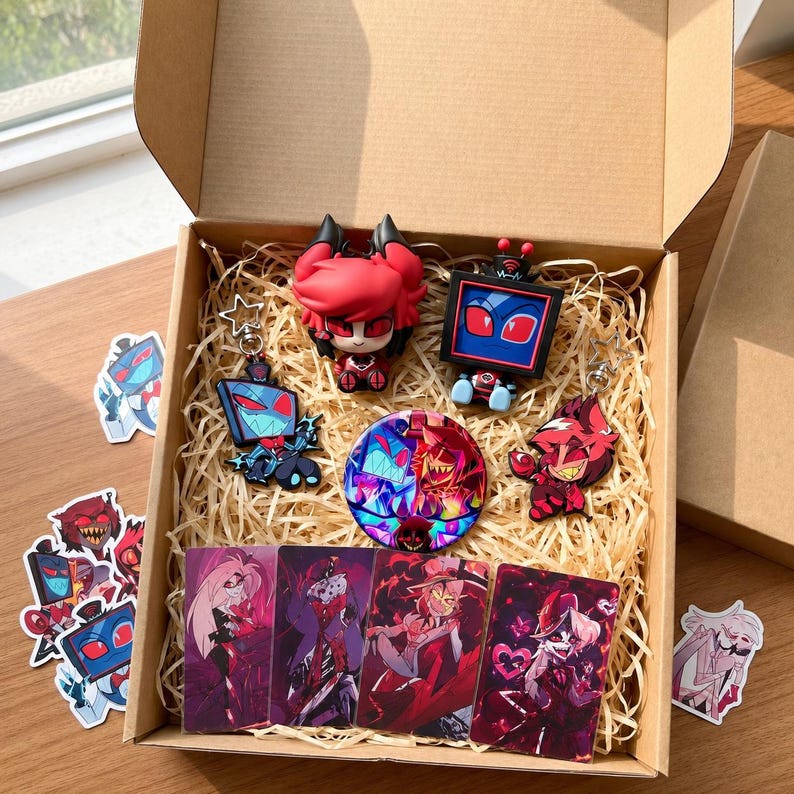 Puede incluir: Una caja de regalo de cart&oacute;n llena de art&iacute;culos de Hazbin Hotel, incluyendo dos figuras de personajes, dos llaveros, un pin circular y cuatro tarjetas art&iacute;sticas rectangulares. Varias pegatinas sueltas est&aacute;n esparcidas alrededor.