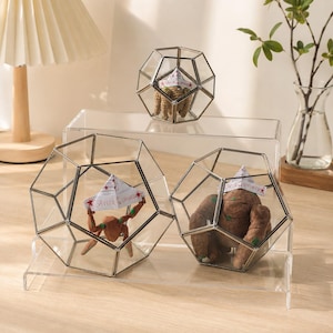 May include: Three geometric glass terrariums with metal frames, each containing a small brown figure wearing a paper hat labeled Savior of the Universe. The terrariums are displayed on a clear acrylic tiered stand, showcasing the figures inside.