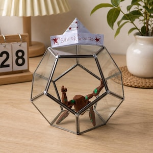 May include: A geometric glass terrarium with a metal frame containing a brown and green angular figurine. A white paper hat sits on top of the glass, featuring red stars and the handwritten text "SAVIOR OF THE UNIVERSE" in black ink.