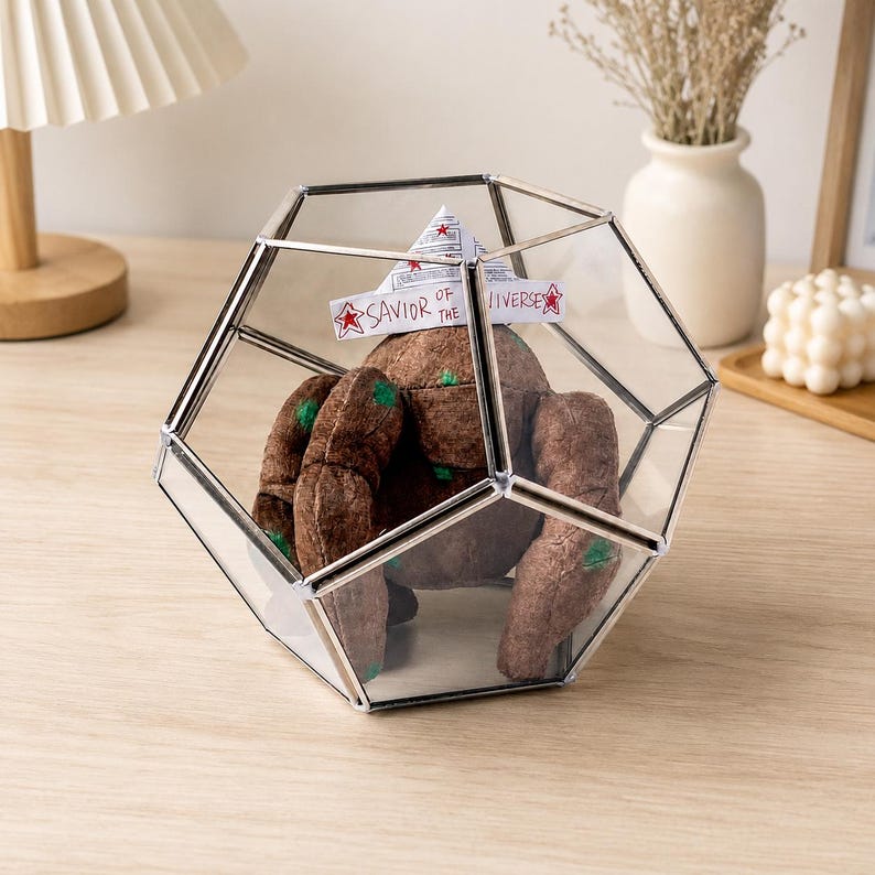 May include: A brown plush creature with green spots sits inside a geometric glass terrarium with silver metal edges. A white paper hat rests on the creature's head, featuring red stars and the handwritten text, Savior of the Universe.