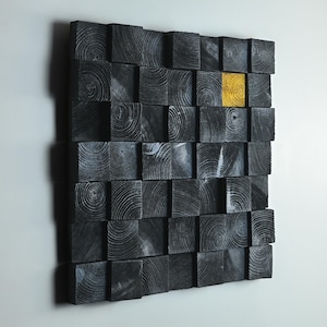 May include: A modern wall art piece composed of numerous dark gray wooden blocks, arranged in a grid pattern. One block is highlighted in a vibrant gold color, creating a striking contrast. The wood grain is visible on each block.