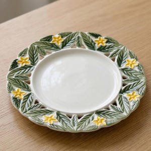 Pode incluir: Um prato de cerâmica branca com uma borda decorativa de folhas verdes e flores amarelas. O prato tem um design circular com uma borda elevada e um acabamento liso e brilhante. O prato está sobre uma superfície de madeira.
