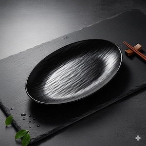 Peut inclure: Un plat de service noir de forme ovale avec une surface texturée, posé sur une ardoise rectangulaire noire. Le plat présente un motif ondulé. Des baguettes reposent sur l'ardoise. L'image a une esthétique sombre et minimaliste.