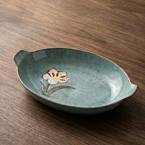 Peut inclure: Un plat de service en céramique de couleur sarcelle avec un motif floral. Le plat a une forme ovale allongée avec de petites poignées. La fleur est blanche avec des accents rouges et jaunes.