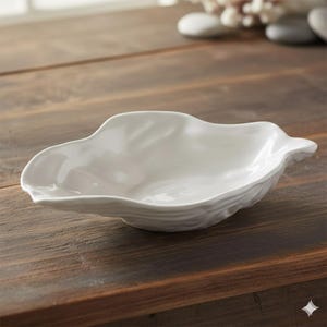 Peut inclure: Un plat de service blanc en forme de feuille. Le plat a un bord ondulé et texturé et un intérieur lisse. Le plat est posé sur une surface en bois foncé. Le plat est vide.