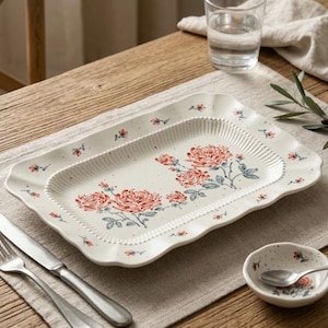Peut inclure: Plat rectangulaire en céramique crème avec un bord festonné. Le plat présente un motif floral de roses rouges et de feuilles vertes. Un petit bol assorti avec une cuillère est à côté du plat. Une fourchette et un couteau sont à gauche.