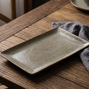 Può includere: Un vassoio rettangolare in ceramica maculata nei toni del grigio e del beige. Il vassoio ha un bordo leggermente rialzato e poggia su un tavolo di legno rustico. Un tovagliolo di lino grigio è parzialmente visibile.