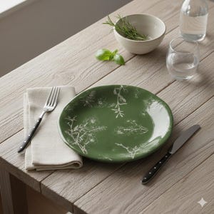 Peut inclure: Une assiette en céramique verte avec des motifs floraux blancs, une fourchette et un couteau à manches noirs sont disposés sur une table en bois clair. Une serviette beige, un petit bol blanc avec des herbes et un verre d'eau sont également présents.