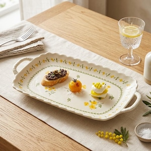 Puede incluir: Una bandeja de servir rectangular de cerámica con bordes festoneados y asas en ambos lados. La bandeja presenta una base de color crema decorada con un delicado patrón de enredaderas verdes y pequeñas flores amarillas. Contiene una tostada, un tomate y queso.