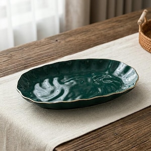 Peut inclure: Un plat de service en céramique vert foncé avec un bord ondulé et un bord doré. Le plat de forme ovale repose sur un chemin de table en lin de couleur neutre, adapté pour servir des entrées ou des desserts.