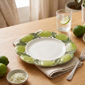 Peut inclure: Assiette en céramique blanche avec une bordure décorative de citrons verts et de feuilles. L'assiette est posée sur une serviette en lin beige sur une table en bois. Un verre d'eau avec une tranche de citron vert, des citrons verts et un petit bol de sel sont également sur la table.