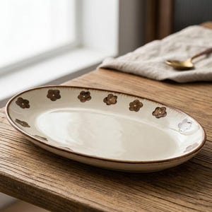 Peut inclure: Un plat de service ovale en céramique avec un intérieur crème et un bord marron. Le plat est décoré de motifs de fleurs marron sur le bord. Le plat est posé sur une surface en bois.
