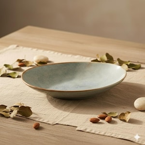 Peut inclure: Un saladier en céramique bleu clair avec un bord marron. Le bol de forme ovale est posé sur une surface en lin beige. Des amandes, de petits cailloux et des feuilles séchées sont dispersés autour du bol. Le bol est probablement utilisé pour servir des aliments.