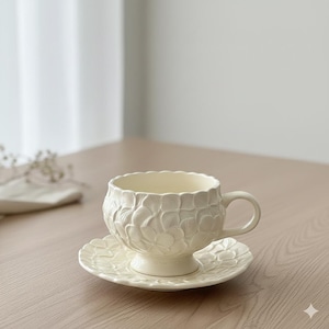 Peut inclure: Service à thé crème composé d'une tasse et d'une soucoupe. La tasse, avec une anse, repose sur un pied. La tasse et la soucoupe présentent un motif floral en relief. L'ensemble est en céramique et posé sur une table en bois.