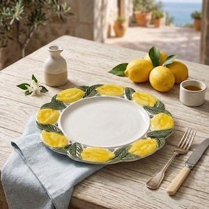 Peut inclure: Une assiette de service en céramique blanche avec un motif de citron sur le bord, posée sur une table en bois avec des citrons, un petit pichet et une tasse d'huile d'olive. Une serviette en lin bleue et des couverts sont également présents.