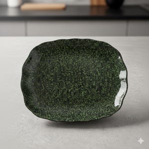 Peut inclure: Une assiette en céramique vert foncé, mouchetée, avec un bord irrégulier et ondulé. L'assiette a une forme légèrement ovale et une surface texturée, adaptée au service des aliments. La couleur de l'assiette est vert forêt foncé avec des mouchetures noires.