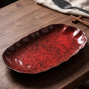 Peut inclure: Un plat de service en céramique rouge et noire mouchetée avec un bord ondulé. Le plat de forme ovale est posé sur une surface en bois, avec un bord foncé et un motif moucheté. Le plat est conçu pour servir des aliments.